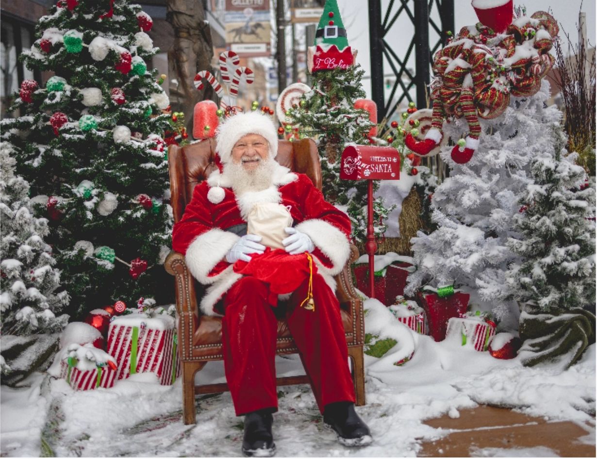 Santa sitting in a chair on a snow-covered sidewalk surrounded by Christmas trees during Plaid Friday.